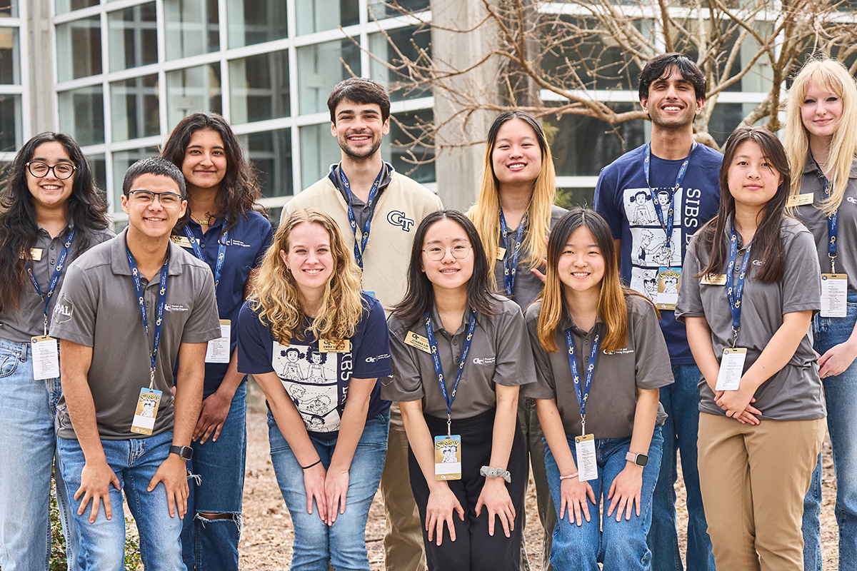 A dozen young men and women stand in two lines facing the camera, smiling brightly